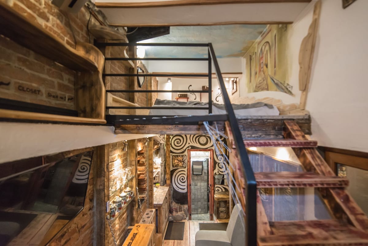 A loft-style living area is presented with a wooden staircase leading to a raised sleeping space. Exposed brick walls are featured, complemented by intricate wall art. The cozy area includes wooden furniture and a small kitchenette, creating an organized and efficient layout.