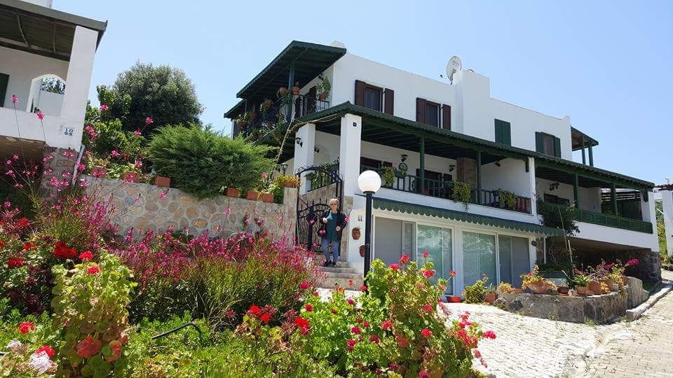 A multi-story building is set amidst lush greenery and colorful flower beds. A stone pathway leads to the entrance, while balconies are adorned with plants. Clear blue skies above complement the charming architecture, creating a serene environment that invites relaxation.