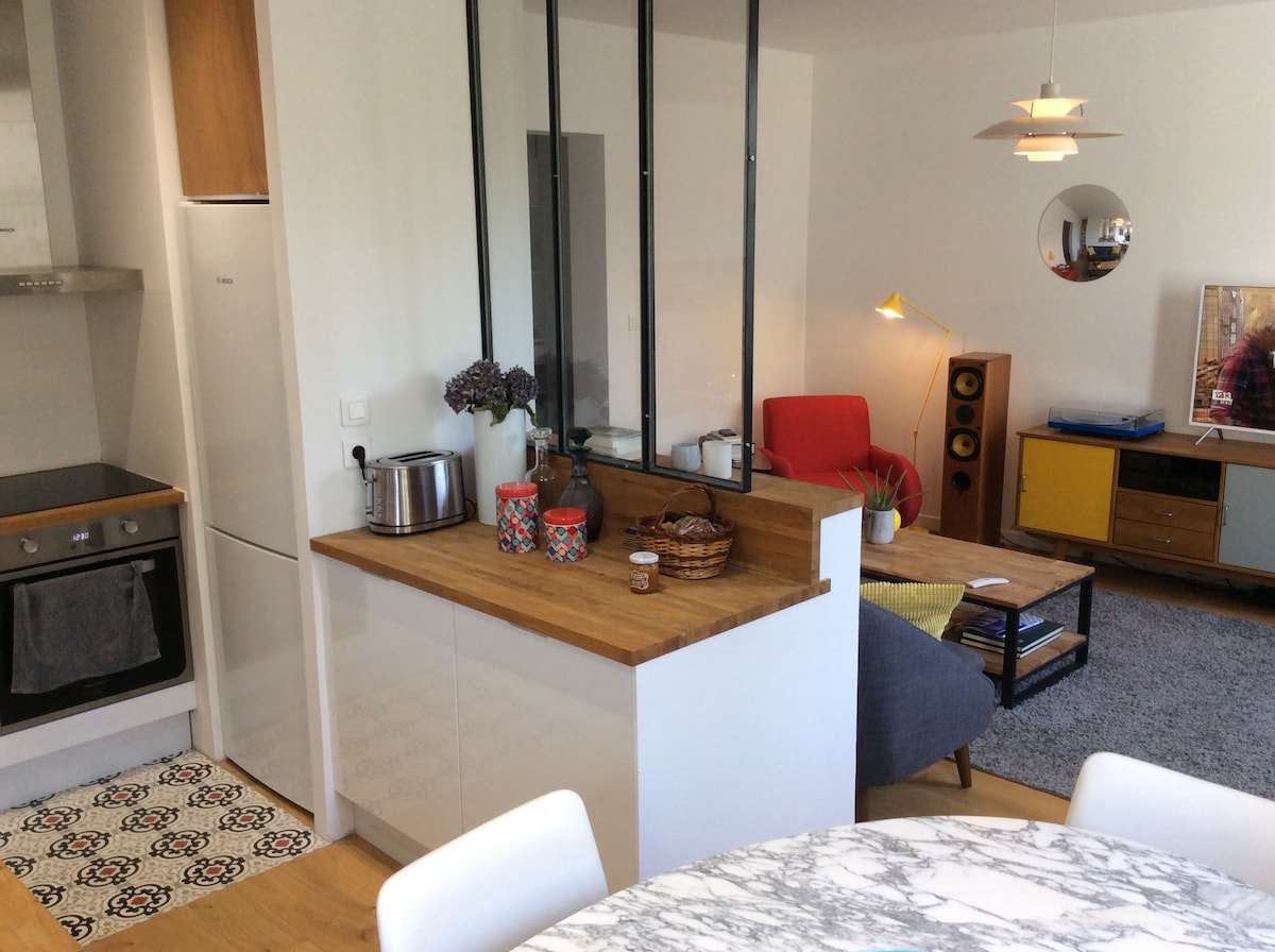 A bright open-plan living space is presented, featuring a modern kitchen with a wooden countertop and stylish cabinets. A cozy seating area is visible, including a red armchair, a gray sofa, and an artfully arranged coffee table. Decorative items and plants add a touch of warmth.