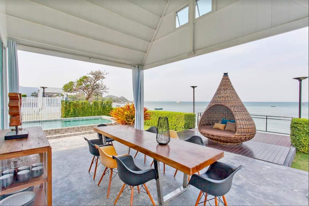 An outdoor dining area features a long wooden table surrounded by comfortable chairs, offering a view of the sea. A hanging egg chair is positioned nearby, while lush greenery and a private pool enhance the atmosphere. The ocean is visible in the background.