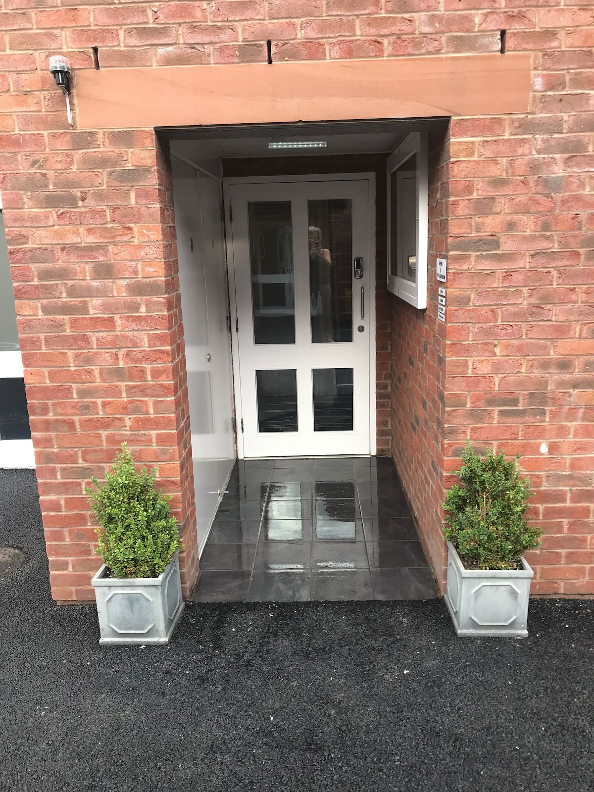 The entrance features a modern, glass-paneled door framed by a brick facade. Planters with decorative greenery are positioned on either side of the entryway, enhancing the welcoming appearance. A smooth, dark tiled surface leads into the building.