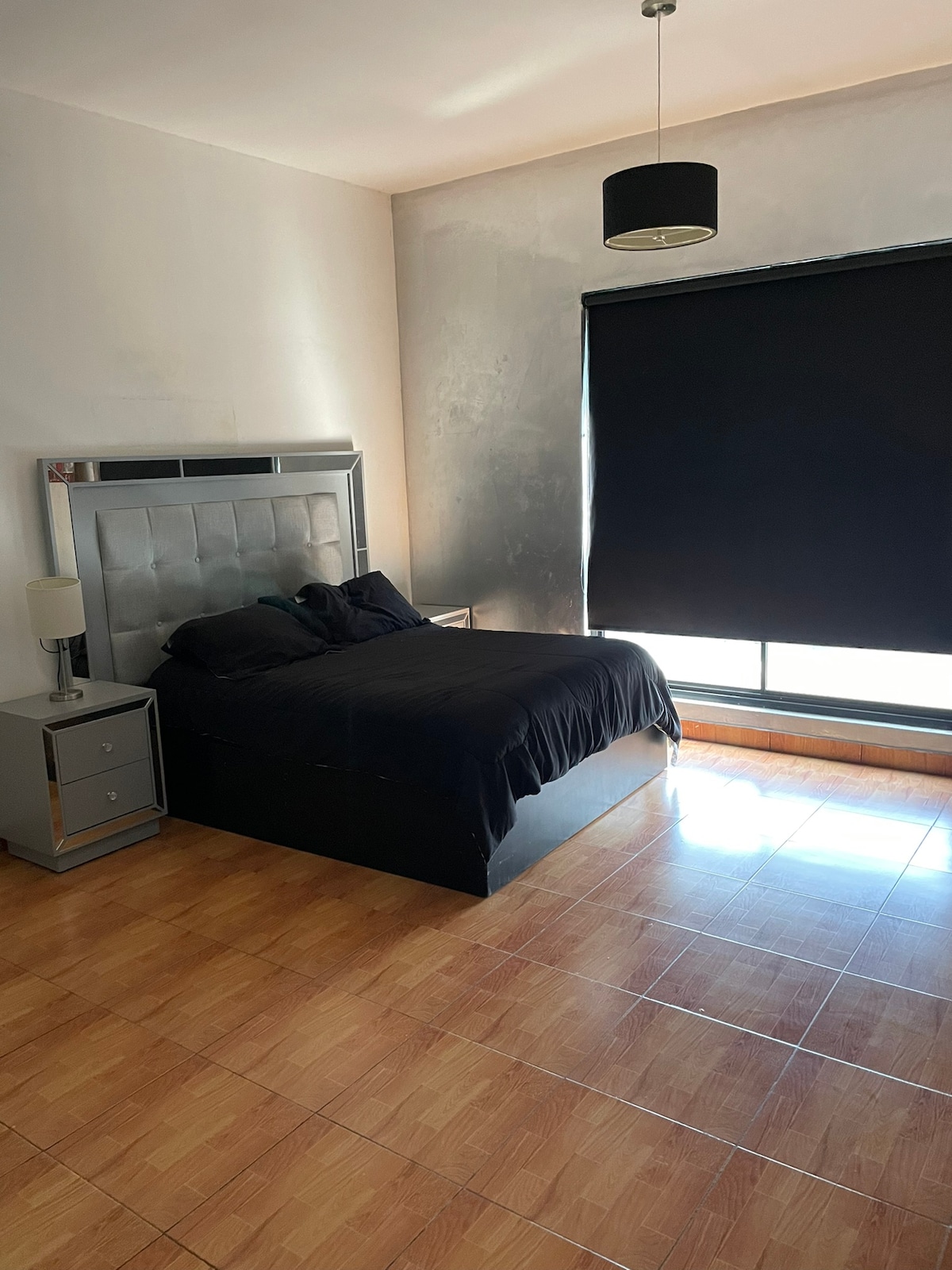 A spacious bedroom features a dark-colored bed with a plush headboard, complemented by two bedside tables with lamps. Large glass windows covered by a black roller shade allow natural light to enter, while terracotta tiles cover the floor.