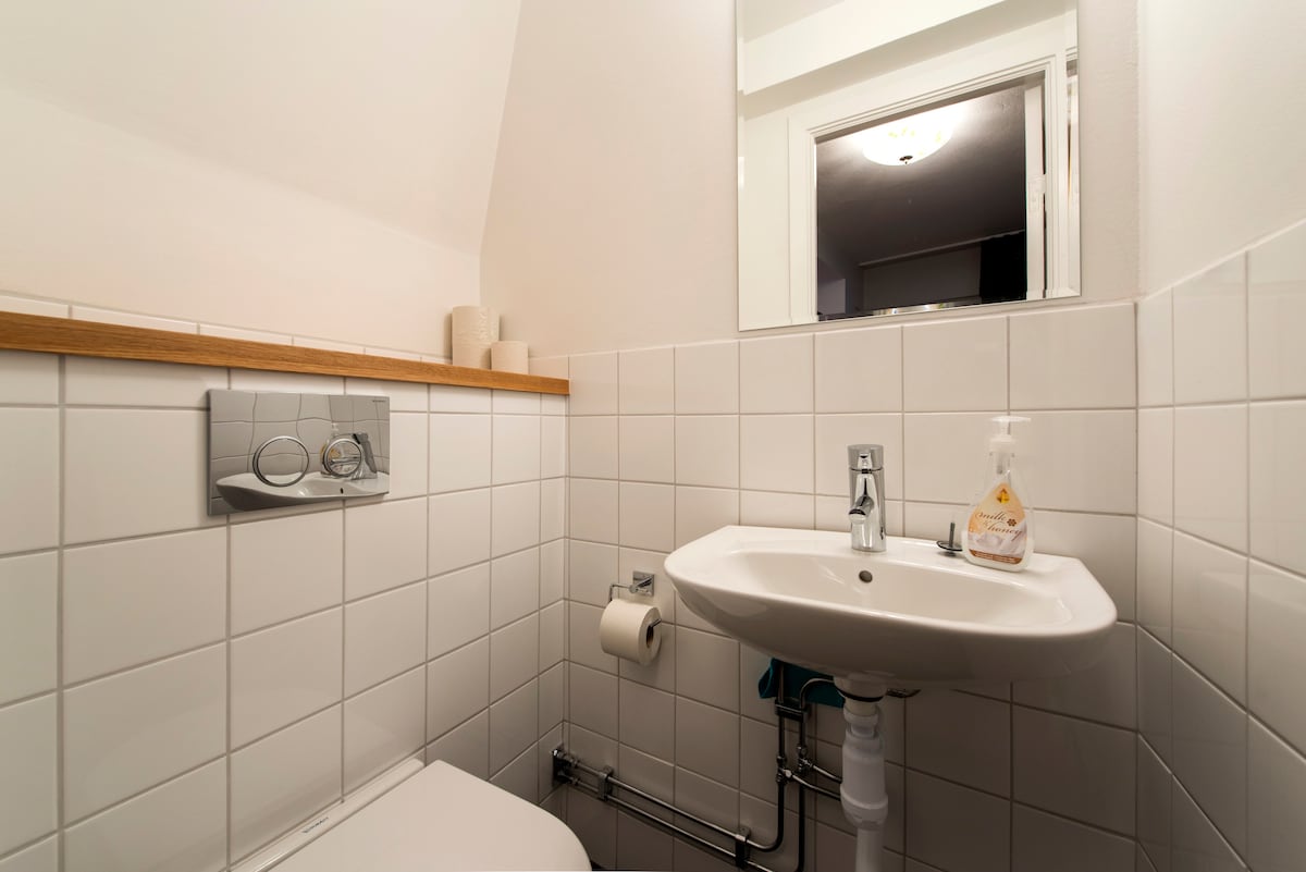 A compact bathroom is showcased, featuring a white tiled wall, a small sink with a mirror above, and essential toiletries. A toilet is positioned next to the sink, and a shelf provides space for additional items. Natural light enhances the clean and modern design.