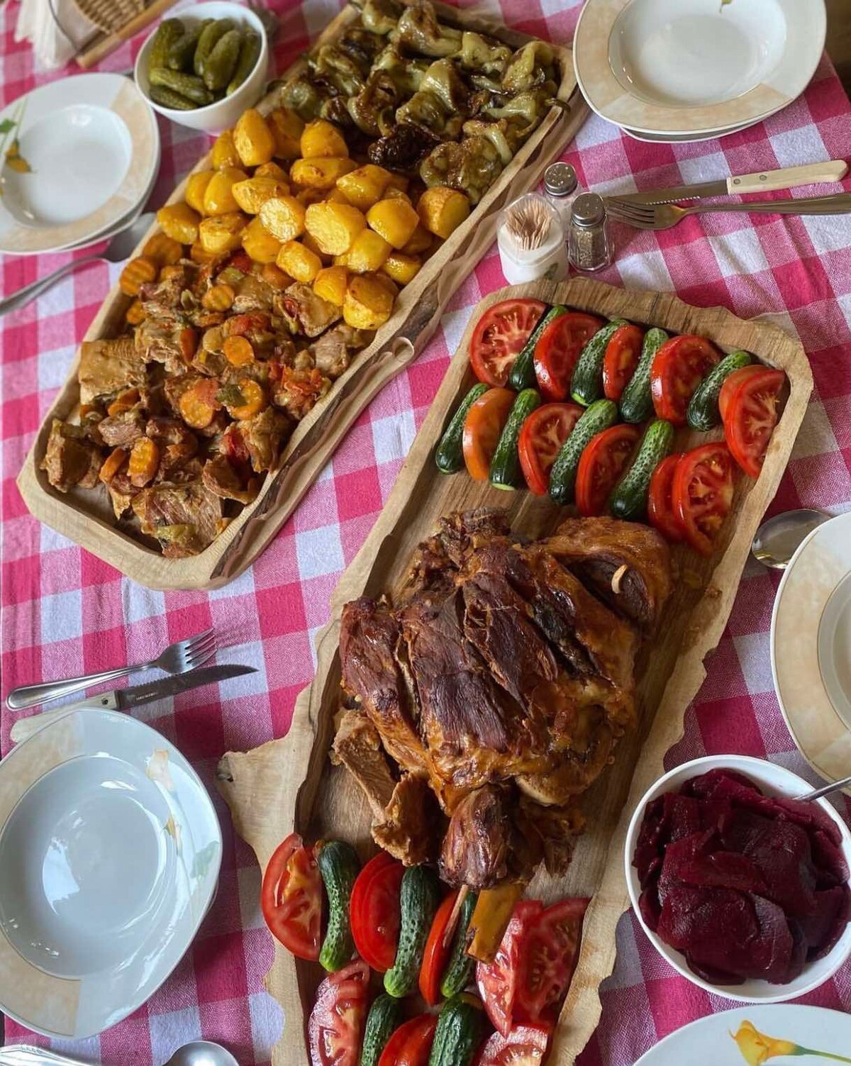 A table set with an abundance of food is displayed. Two large wooden platters hold roasted meats, seasoned vegetables, and fresh salads, featuring vibrant tomatoes and cucumbers. Plates are arranged neatly, complemented by utensils and condiments, set against a checkered tablecloth.