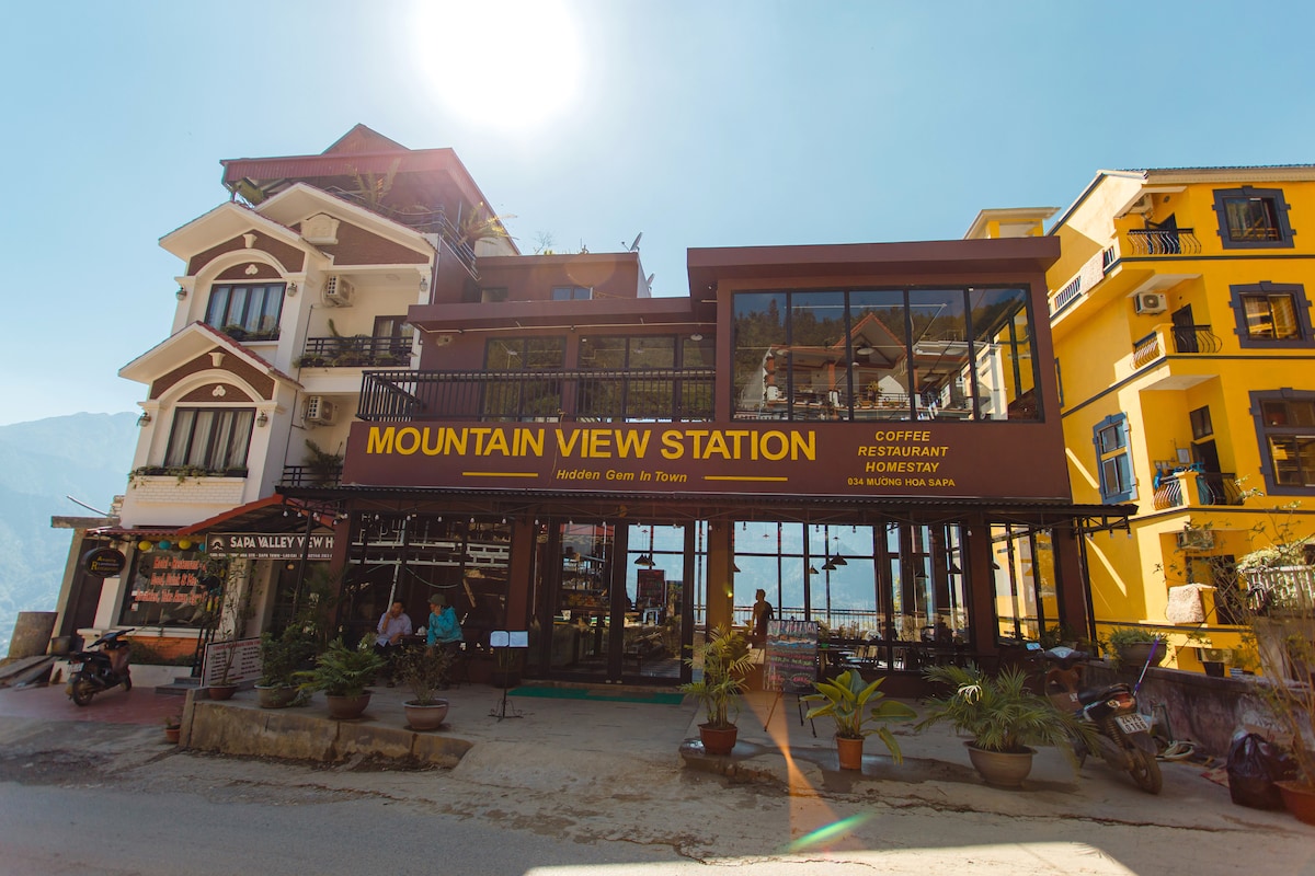 The image showcases the Mountain View Station, a multi-level building featuring a restaurant with large glass windows offering panoramic views. The entrance is adorned with potted plants, and the exterior displays welcoming signage. Surrounding mountains are faintly visible in the background.