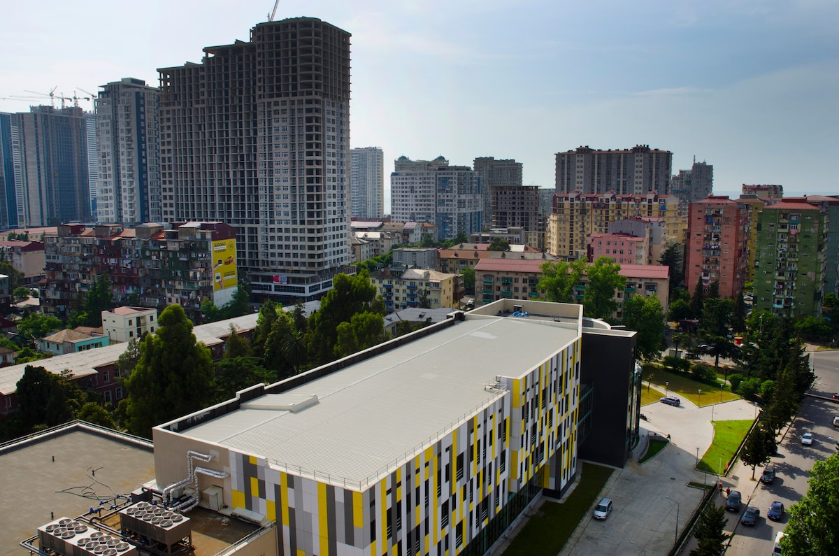 The image displays a modern residential building featuring a colorful facade, nestled among a diverse mix of high-rise and low-rise structures. A clear sky serves as a backdrop, with urban developments and greenery visible in the surrounding area.