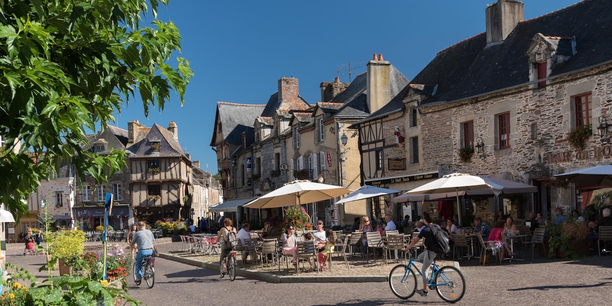 A charming town square is captured under a clear blue sky, with historic buildings lining the perimeter. Outdoor seating areas are filled with patrons enjoying their meals, while a cyclist passes through. Umbrellas provide shade over tables adorned with flowers, fostering a lively atmosphere.