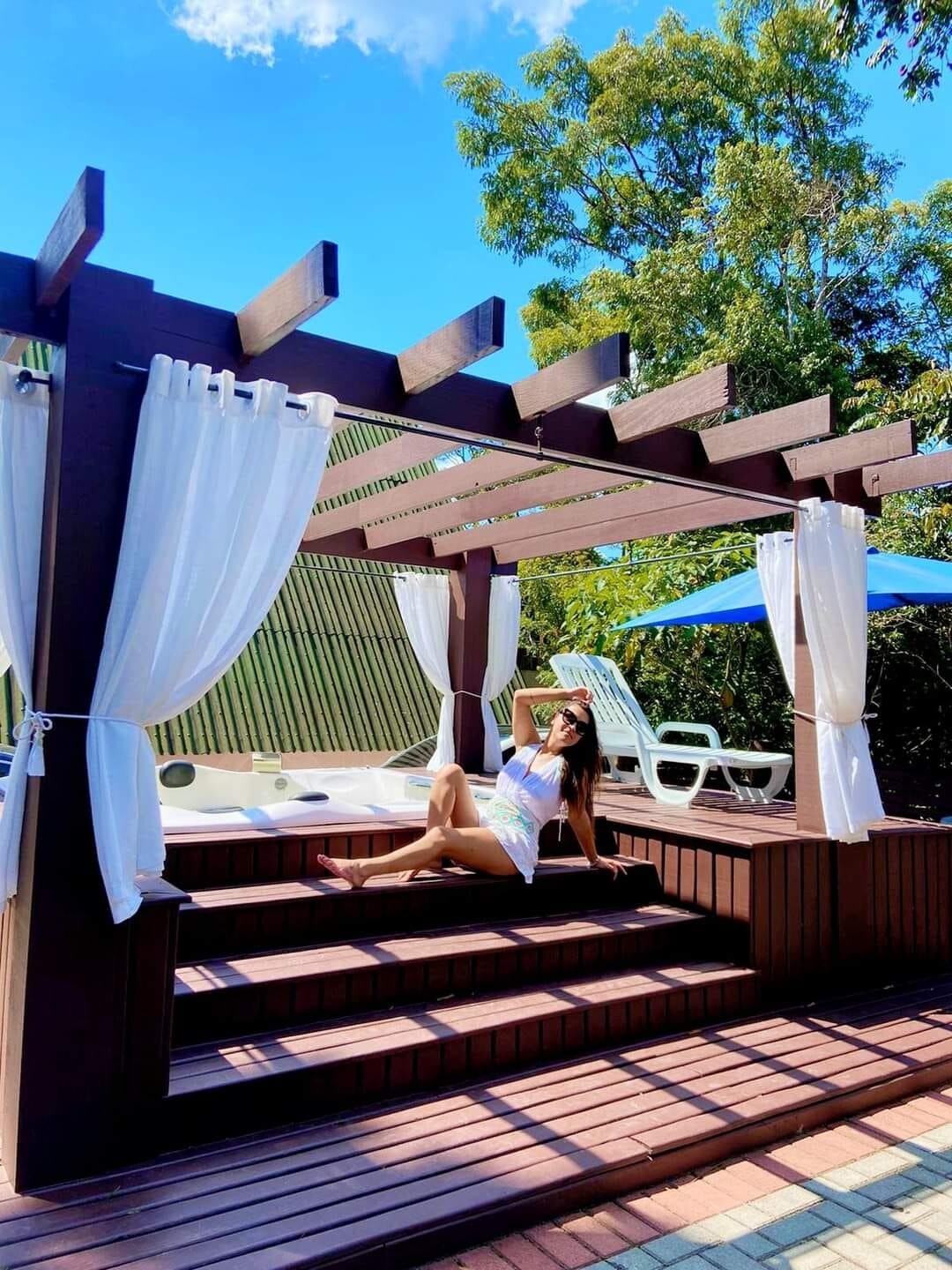 An outdoor area features a wooden deck with a shaded pergola, adorned with flowing white curtains. A spacious jacuzzi is visible in the background, surrounded by lounging chairs under blue umbrellas. Lush greenery complements the relaxing atmosphere.
