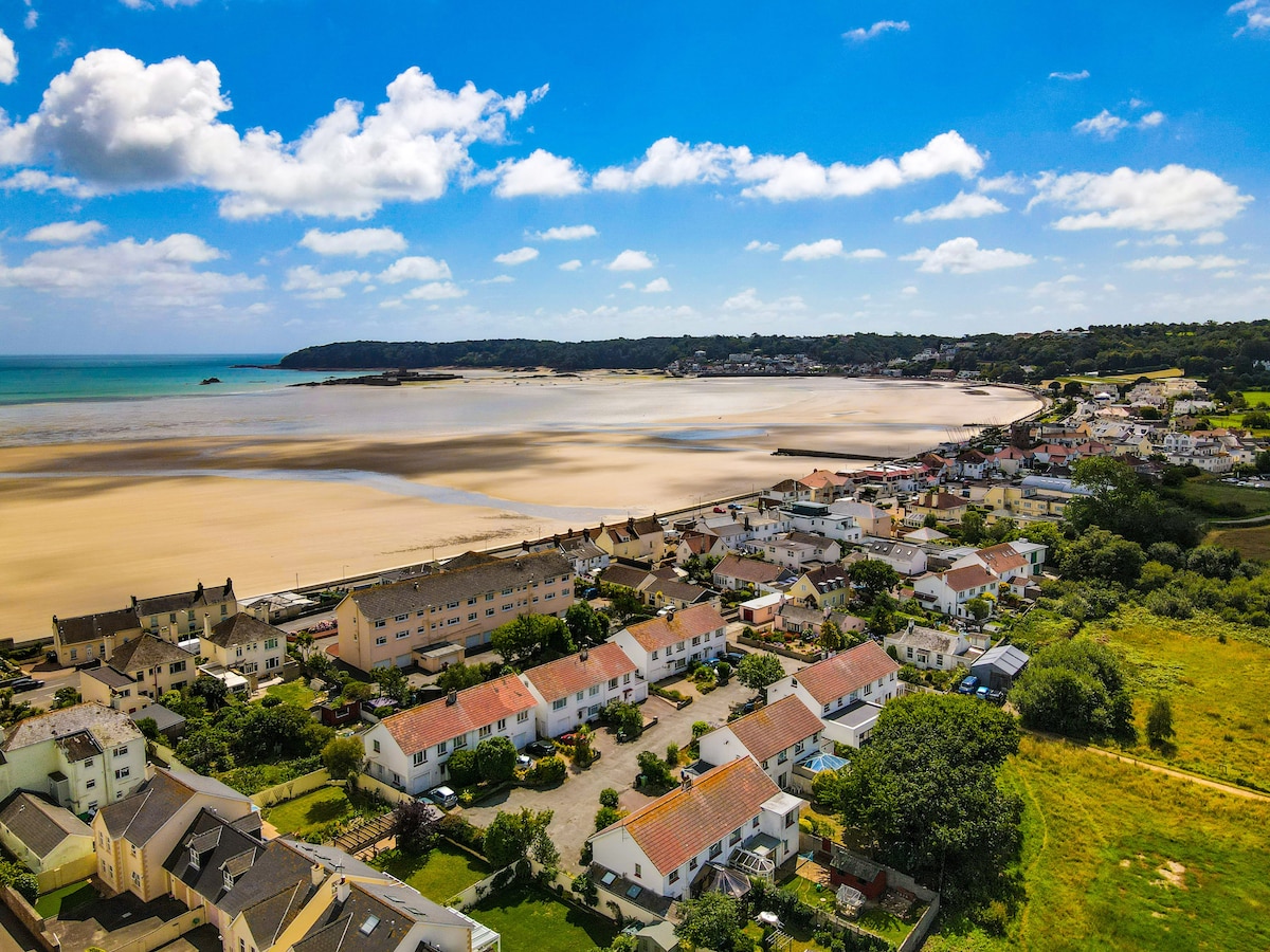 An aerial view captures a coastal landscape, featuring sandy shores and gentle waves. Residential homes line the beach, showcasing a variety of architectural styles. Lush green hills rise in the background, with the expanse of blue sky dotted with fluffy clouds overhead.