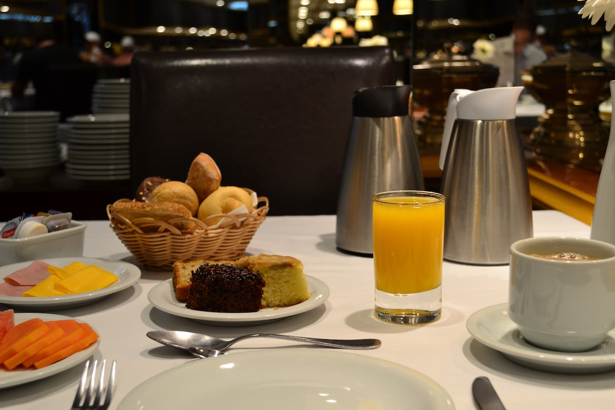 A breakfast table is set with a white tablecloth. A woven basket holds an assortment of baked goods. Plates feature sliced fruits, pastries, and a yellow beverage. A coffee cup is positioned alongside two silver containers, offering a selection of condiments.