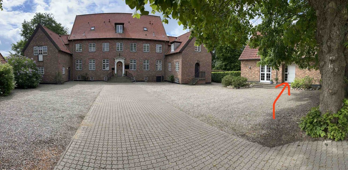 A spacious courtyard is framed by a brick building with multiple stories and gabled roofs. A well-maintained path of pavers leads to an entrance visible on the right, with lush greenery surrounding the area and providing a serene environment.
