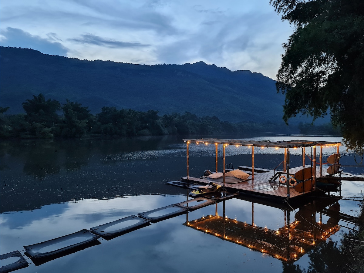 A serene riverside raft is illuminated by soft string lights, reflecting gently on the water's surface. Lush mountains frame the background, while a few comfortable chairs invite relaxation. The peaceful atmosphere is enhanced by the tranquil nature surrounding the area.