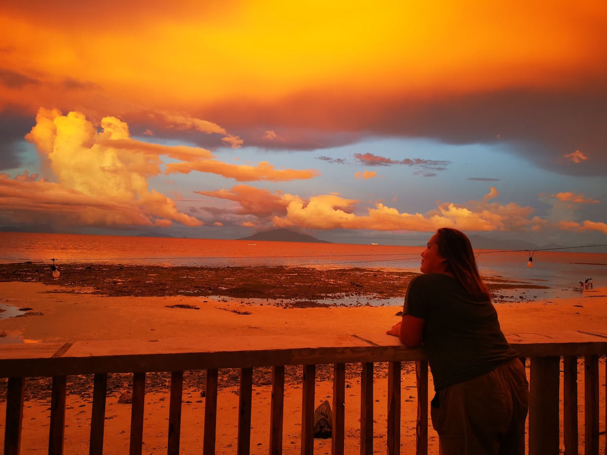 A serene beach vista is captured during sunset, showcasing a colorful sky with vibrant orange and blue hues. The shoreline is visible in the foreground, while gentle waves reflect the evening light. A railing in the foreground frames the scene, inviting moments of reflection.
