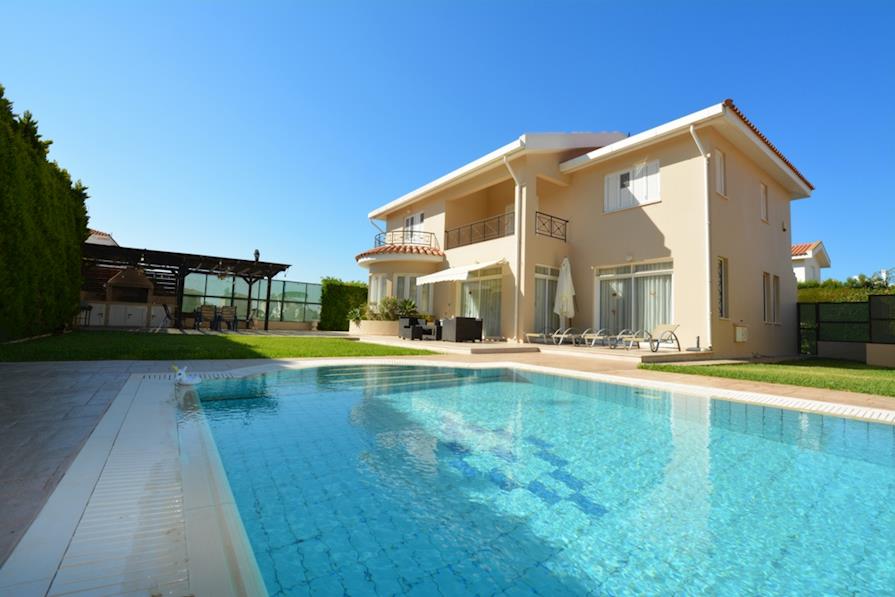 The exterior of the villa is presented with a vast private pool, reflecting clear blue skies. A spacious patio area includes seating arrangements and a shaded outdoor structure. Lush green grass surrounds the pool, enhancing the serene outdoor space.