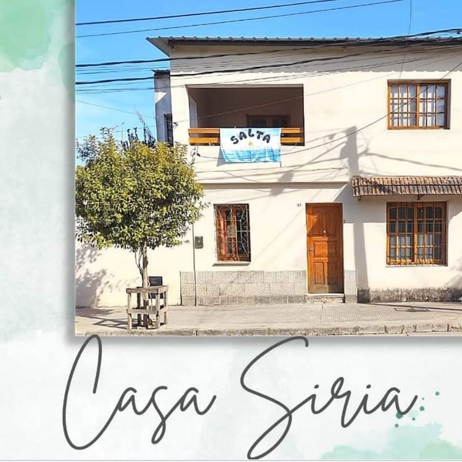The exterior of Casa Siria displays a two-story structure with a welcoming wooden door and large windows. A banner with 'Salta' is prominently displayed on the balcony. A small tree adds a touch of greenery to the entrance, and a simplistic table is visible by the walkway.