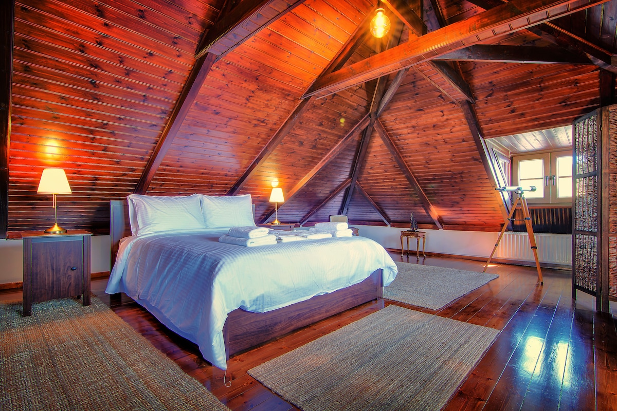 A cozy attic room features a spacious bed with crisp white linens and soft towels arranged neatly. The room is illuminated by warm lamps and showcases wooden beams and flooring, creating a rustic atmosphere. A telescope is positioned near a window, offering a view of the surroundings.