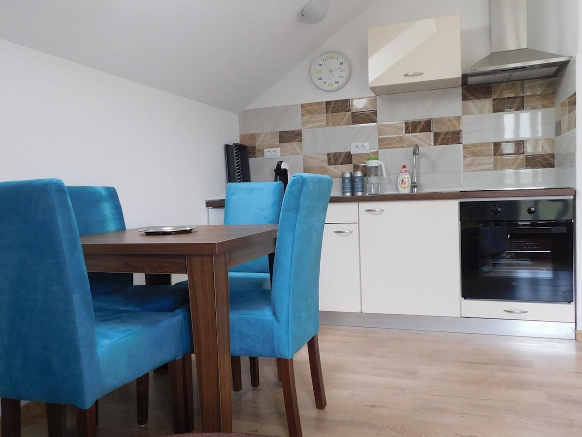 A compact kitchen area features a table with four blue chairs, creating a vibrant dining space. The wall is decorated with a modern tile backsplash. Basic kitchen appliances, including an oven and a coffee maker, are visible, alongside countertop essentials and a wall clock.