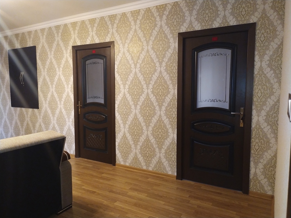 A hallway features two elegant dark wood doors with decorative glass panels, framed by a classic patterned wallpaper in warm tones. A portion of a comfortable couch is visible on the left, complementing the wooden flooring.
