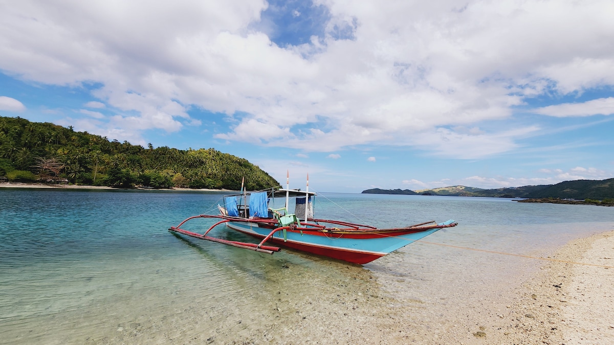 A traditional boat with a colorful hull is anchored in clear, shallow waters. The sandy beach extends along the shoreline, bordered by lush green hills under a sky dotted with clouds, creating a tranquil coastal atmosphere.