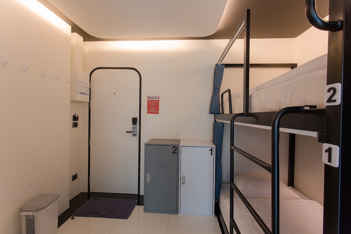 A compact dormitory room is equipped with a bunk bed featuring two sleeping areas. Adjacent to the bed, two storage cabinets are placed against the wall. The room includes a door with a notice and hooks for belongings. A waste bin is positioned on the floor.