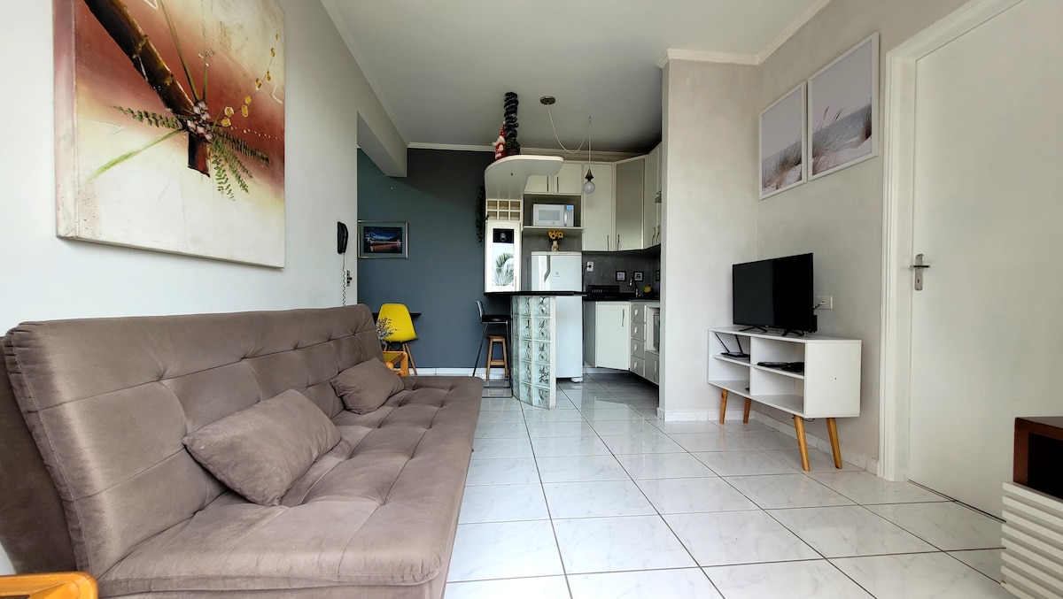 A modern living area features a comfortable sofa bed positioned against a wall adorned with artwork. The light-colored tile floor is complemented by a minimalist TV unit and contemporary kitchen area, while natural light enters through windows, enhancing the inviting space.