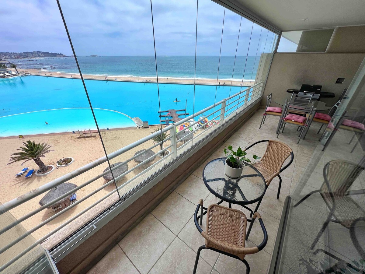 A glass-enclosed balcony provides an unobstructed view of the turquoise waters and sandy beach below. A small round table with two chairs sits adjacent to a seating area equipped with a gas grill. Light-colored tile flooring complements the space.