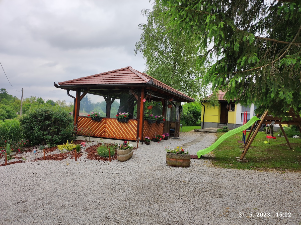 A spacious outdoor area features a wooden gazebo adorned with potted flowers, offering shade. Nearby, a green slide is positioned near a playset, surrounded by gravel pathways and landscaped gardens. The home in the background complements the serene setting of greenery and open space.