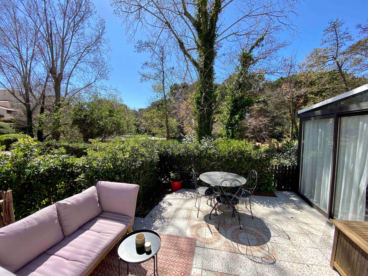 A sunny terrace features a light pink sofa and a round dining table with metal chairs. Lush greenery surrounds the area, providing a serene view of trees and shrubs. A small planter adds a touch of color, while natural light brightens the space.