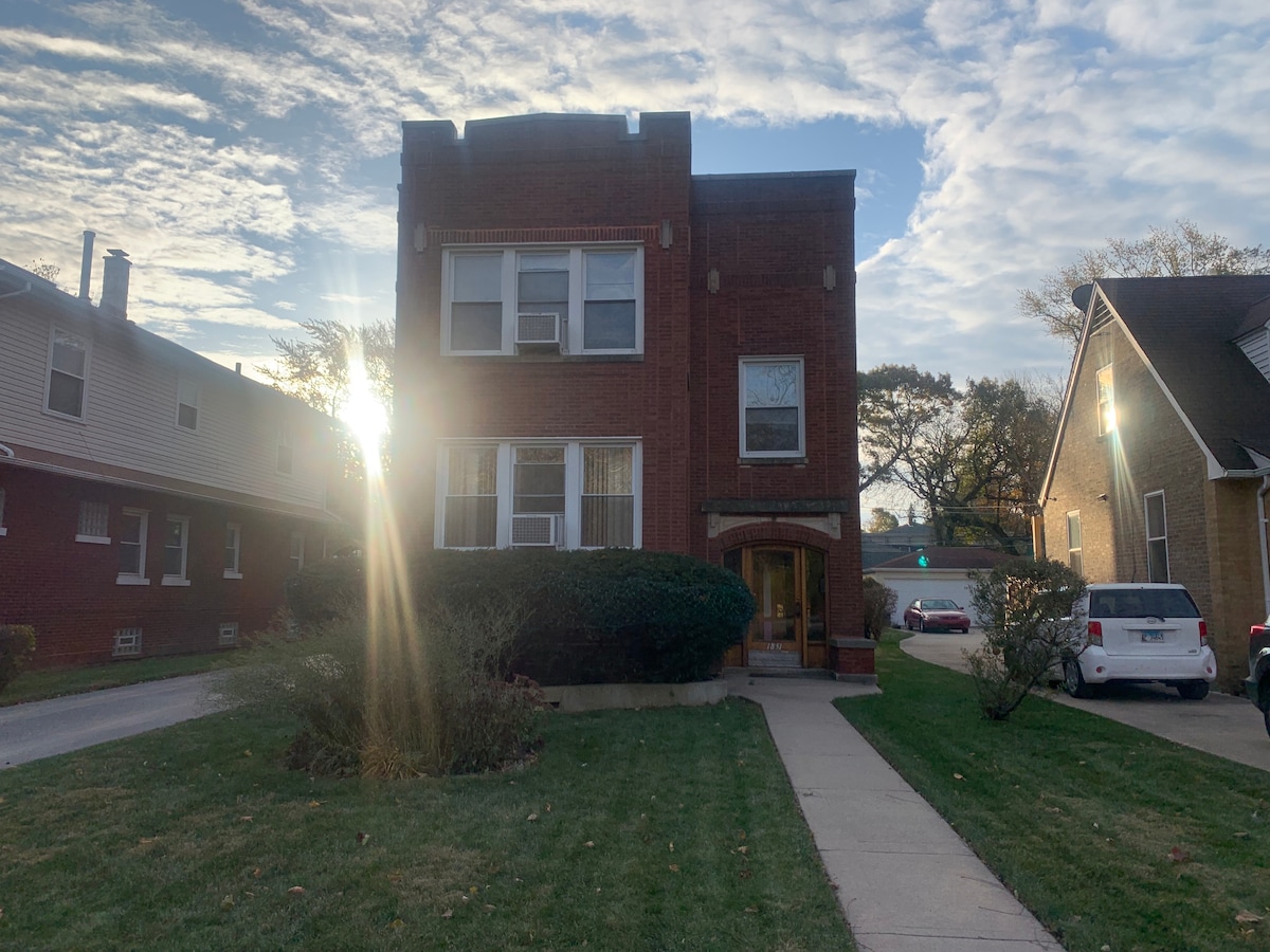 A well-maintained red brick building is depicted, featuring multiple windows that allow natural light to filter through. The front yard is landscaped with green grass and a pathway leading to the entrance. The sky is partly cloudy, creating a serene backdrop.