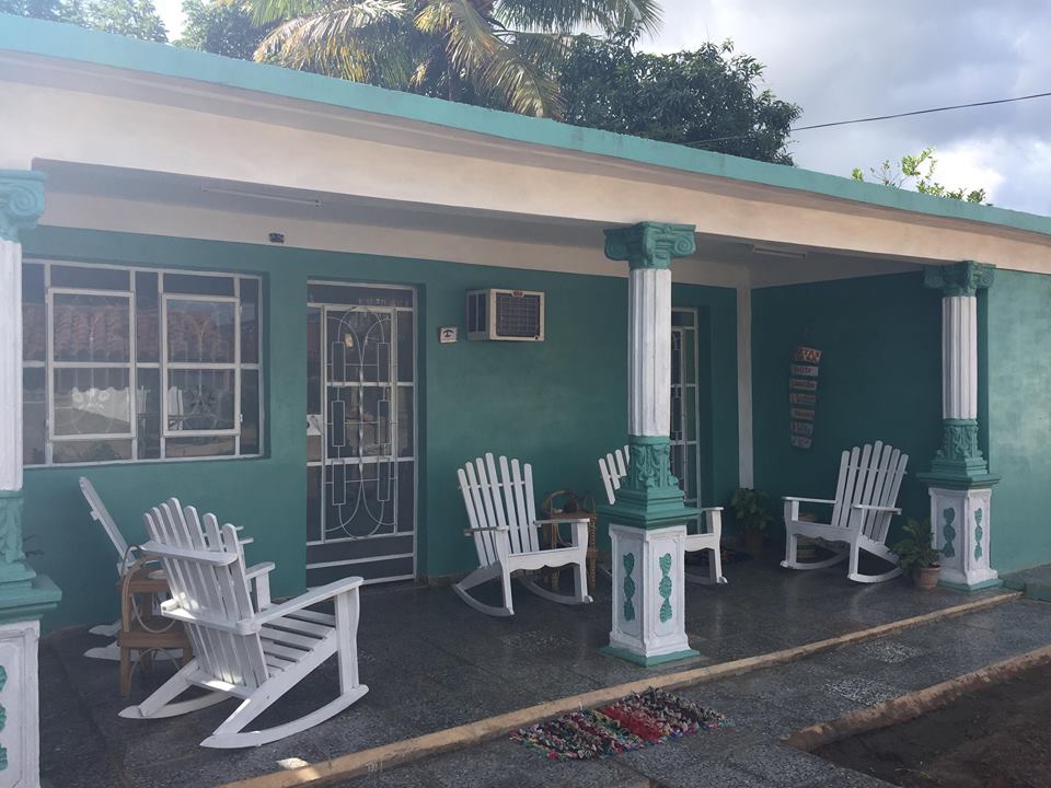 A welcoming porch features several white rocking chairs arranged around a central space, inviting relaxation. The walls are painted in a soothing shade of teal, complemented by decorative columns. A window and a door provide access to the interior, with an air conditioning unit placed above.