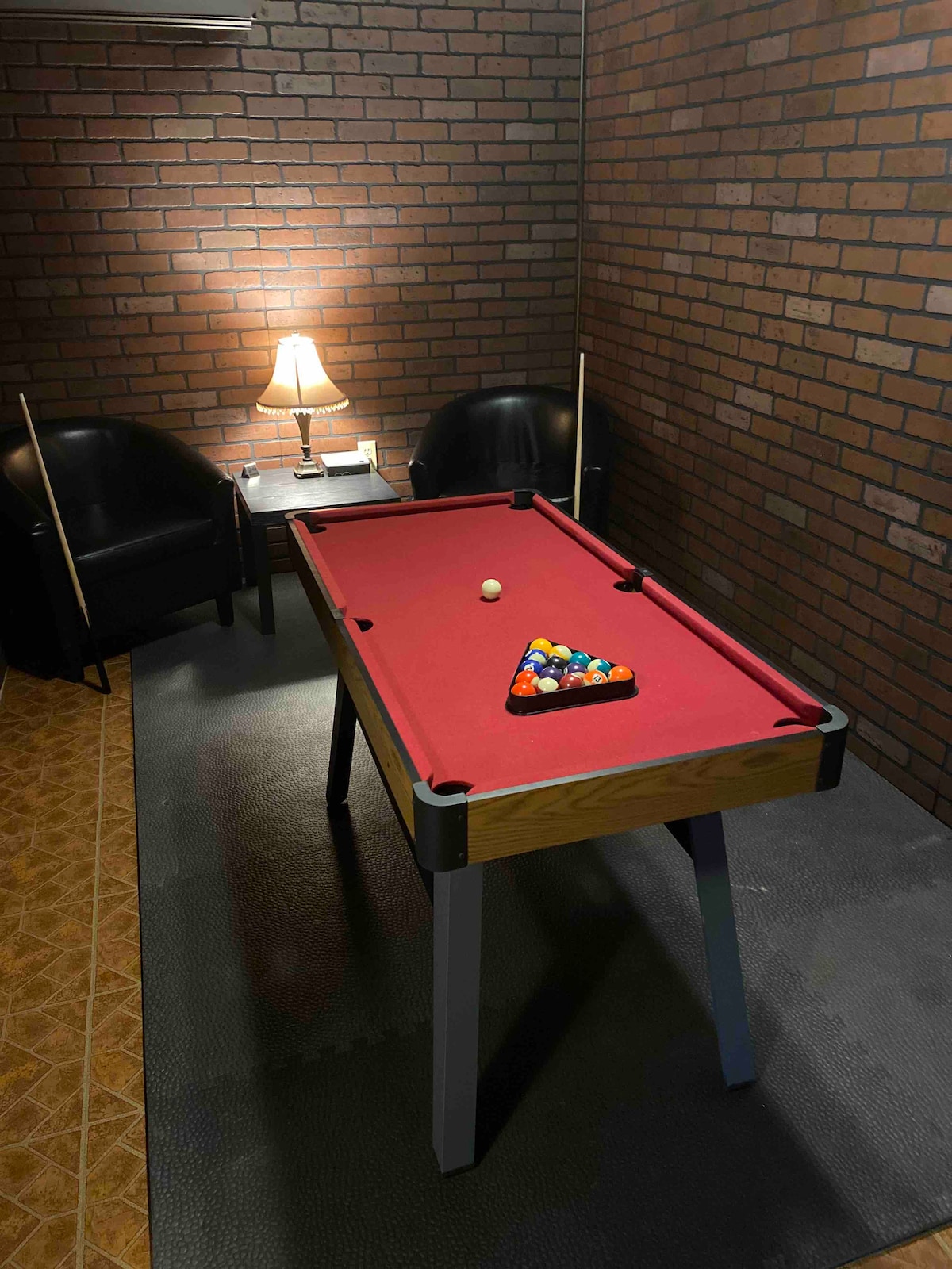 A pool table is set in a cozy corner, surrounded by two black armchairs and a lamp providing soft light. The rich brown brick wall adds warmth to the space, and a triangle of balls is neatly arranged on the table, ready for play.