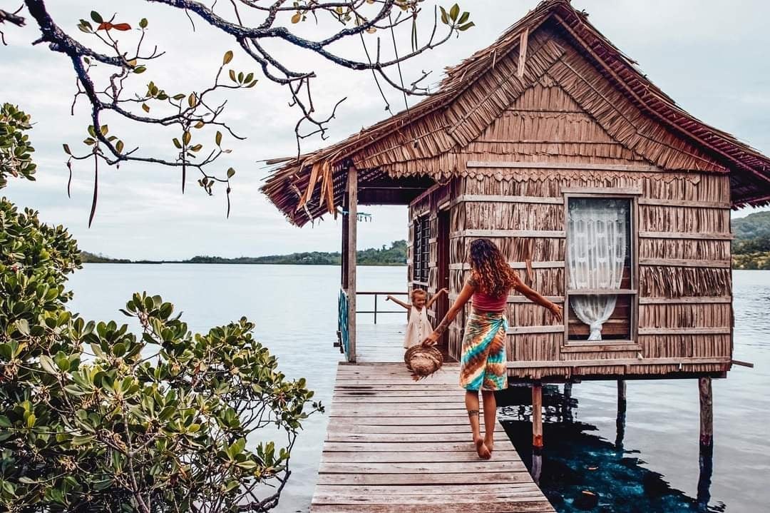 A wooden bungalow with a thatched roof is positioned on stilts over calm water, surrounded by lush greenery. A wooden pathway leads to the entry, while a window with sheer curtains adds to the serene setting. The sky is overcast, reflecting a tranquil atmosphere.