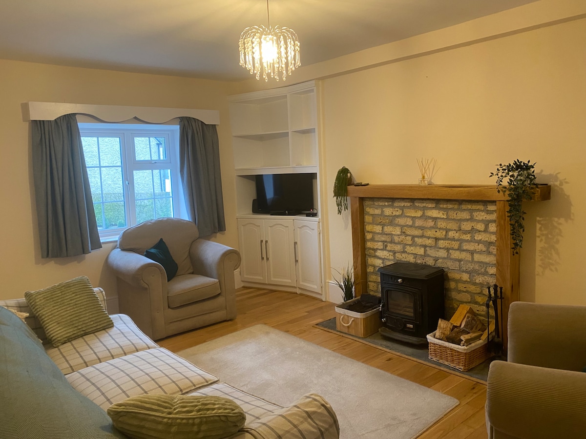 A cozy sitting room features a comfortable couch and a plush armchair, complemented by a wooden coffee table. A wood-burning stove is set within a charming stone fireplace, with a decorative plant and a basket for logs nearby. Soft lighting enhances the inviting atmosphere.