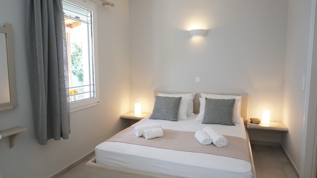 A bright sleeping area features a spacious bed with soft bedding and two accent pillows. Light from a nearby window brightens the room, with soft-gray curtains partially drawn. Two illuminated bedside fixtures provide gentle lighting, and a simple mirror is mounted on the wall.