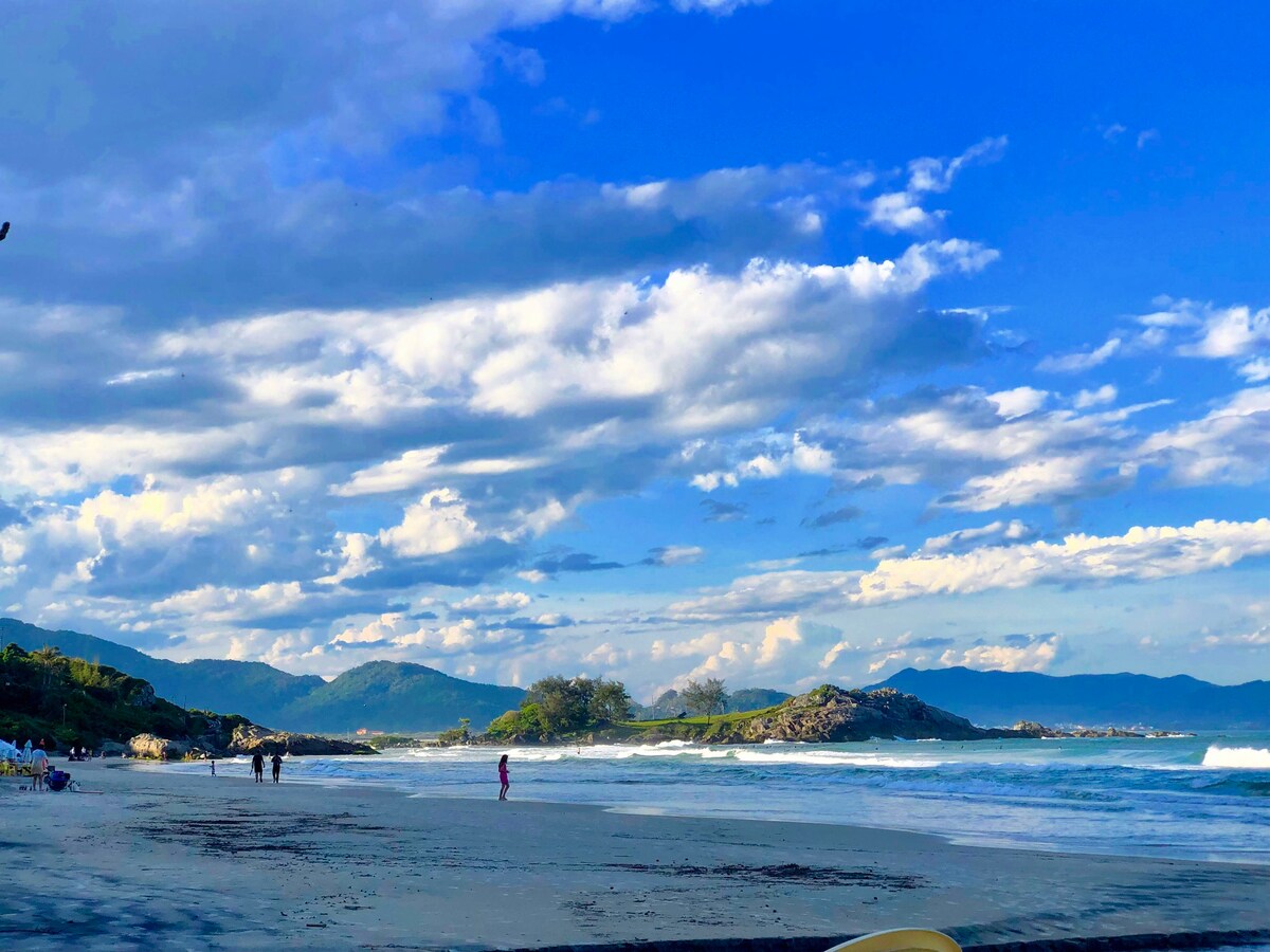 A serene beach scene is captured under a vast sky filled with scattered clouds. Gentle waves lap against the shore, where a few individuals stroll along the water's edge. Lush greenery and distant hills frame the background, offering a sense of tranquility and natural beauty.