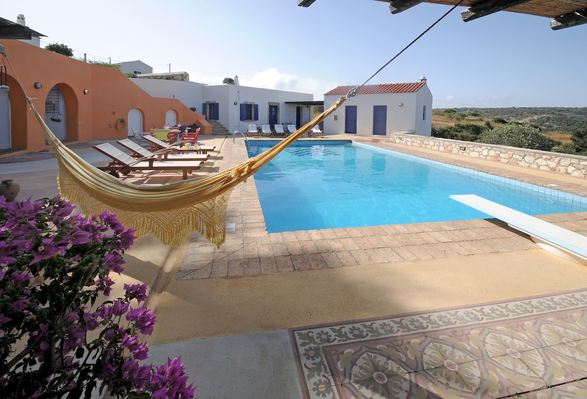 A spacious outdoor pool area features a clear blue swimming pool surrounded by sun loungers and vibrant flowers. A unique hammock adds a touch of relaxation, while nearby buildings are designed in a traditional style, blending seamlessly with the tranquil landscape.