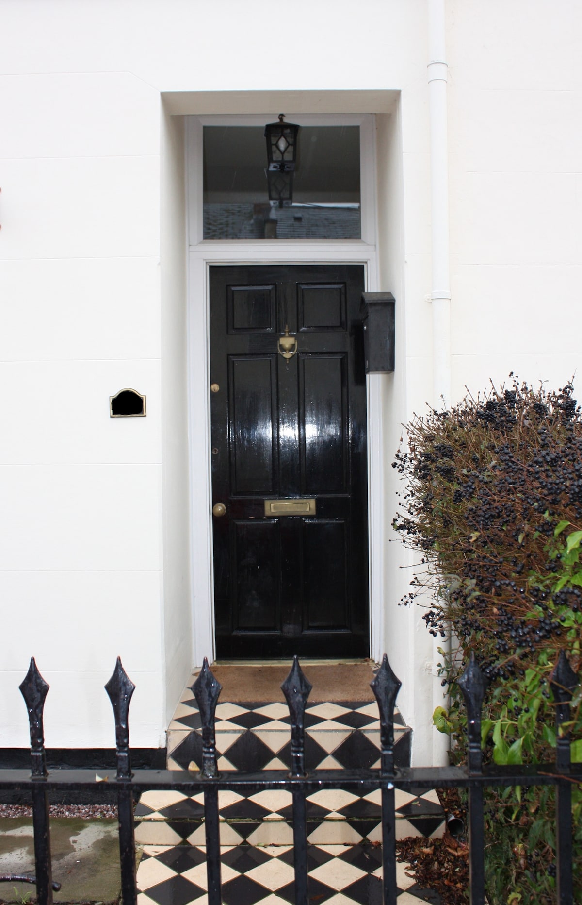 A classic black front door is framed by white walls, featuring a brass door knocker and mail slot. The entrance is approached by a patterned checkered path, lined by a low iron fence with pointed tops. Lush greenery borders the entryway.