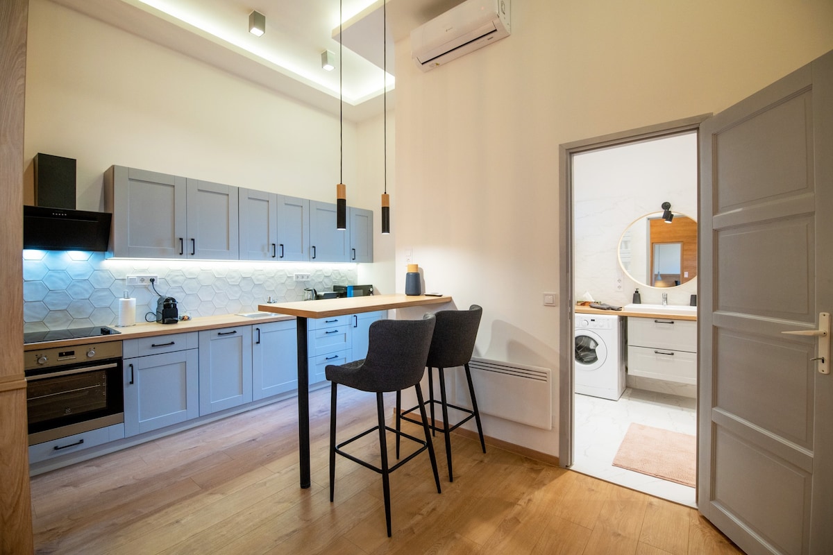 A modern kitchen area features a spacious countertop with stylish bar stools. Equipped with contemporary appliances including an oven and dishwasher, the kitchen is well-lit with under-cabinet lighting. A door leads to a bright bathroom, enhancing the open layout.