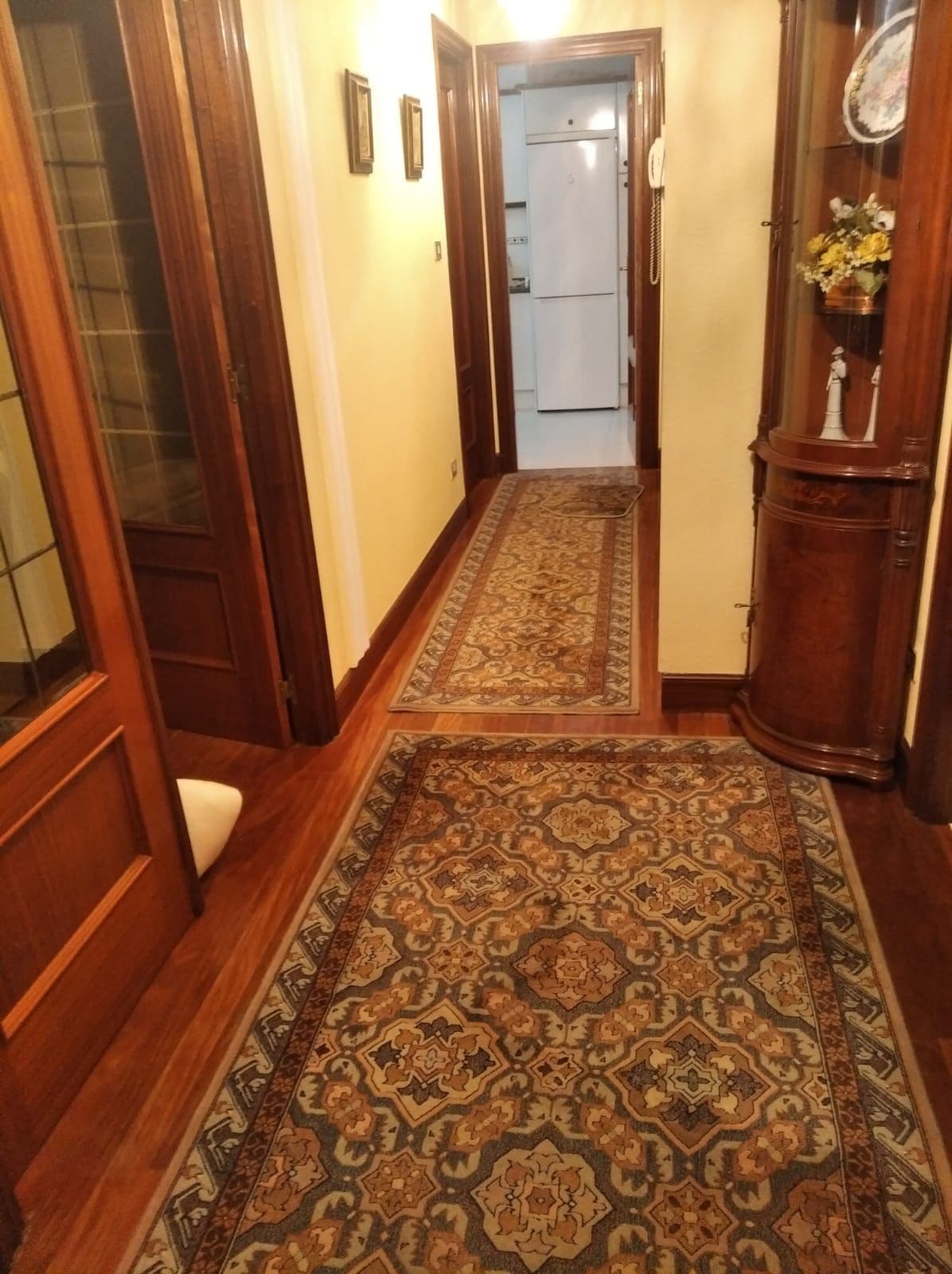 A long hallway is lined with intricate patterned carpets, enhancing the warm tones of the wooden paneling. Doorways to rooms are visible on either side, while a display cabinet stands at one end, featuring decorative items. Soft lighting highlights the inviting atmosphere of the corridor.