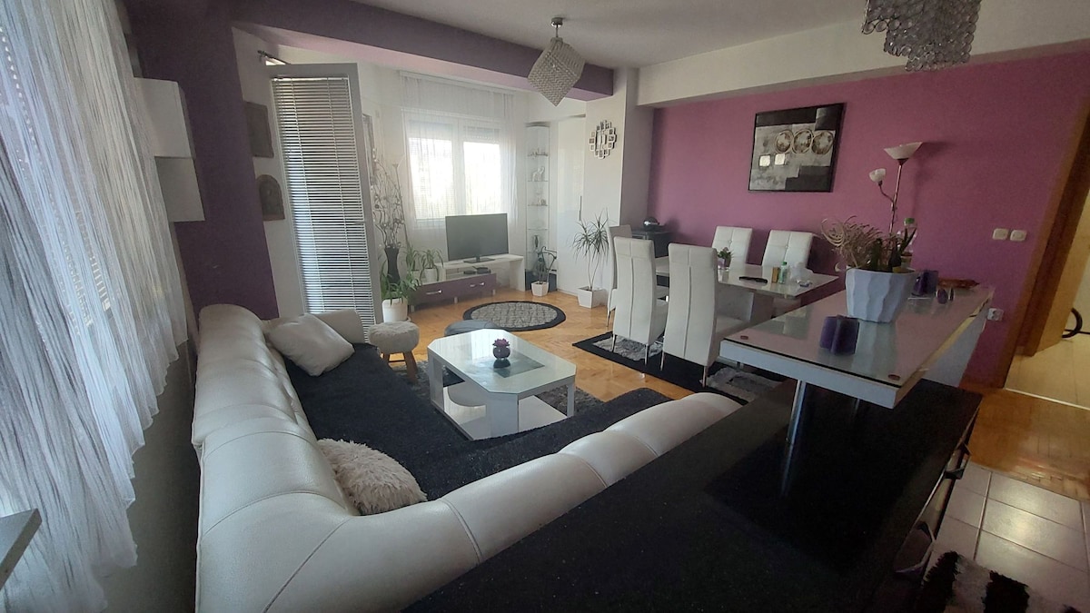A spacious living area is arranged with a large white couch and a coffee table at its center. A dining table with white chairs sits adjacent, while the space is illuminated by natural light from large windows and accented with indoor plants.