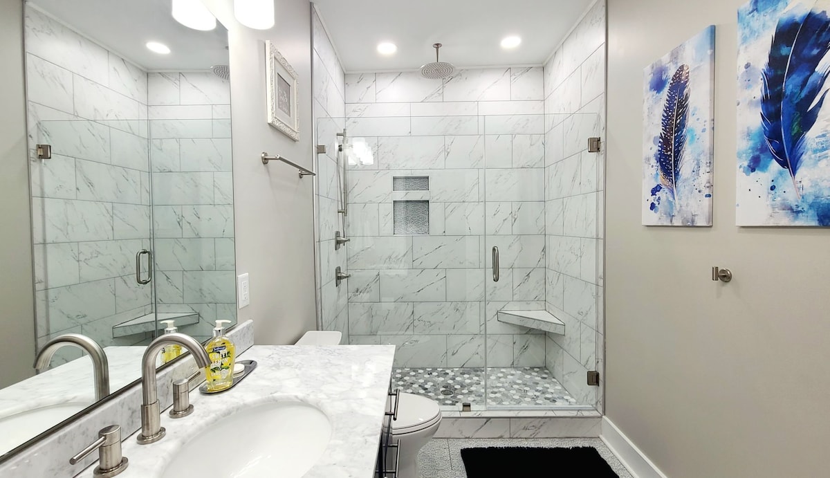 A modern bathroom features a spacious walk-in shower enclosed by glass panels. Elegant marble tiles line the walls and floor, complemented by a marble vanity with dual sinks. The shower includes a rainfall showerhead and a built-in bench. Two artistic pieces adorn the walls.