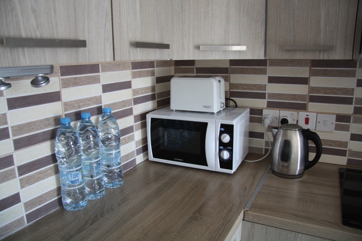 A clean kitchen space features a microwave and a kettle positioned on a countertop. Three bottles of water are placed nearby, complemented by a toaster. The cabinetry has a light wood finish, and a patterned backsplash adds visual interest.
