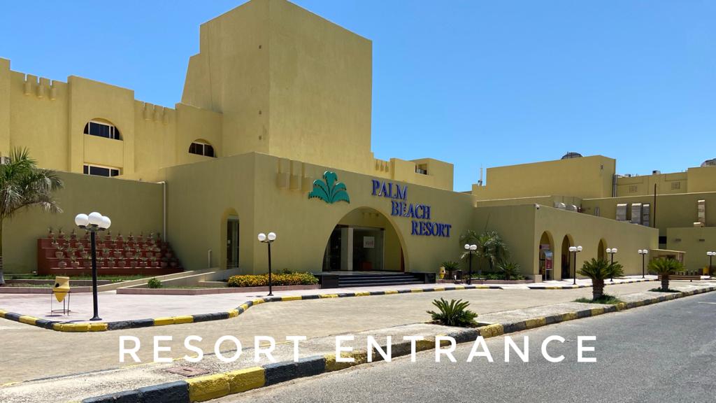 The entrance of Palm Beach Resort is highlighted by the building's distinctive yellow façade and the prominent 'Palm Beach Resort' sign. A spacious pathway leads to the main entrance, flanked by palm trees and decorative lighting, with a clear blue sky above.