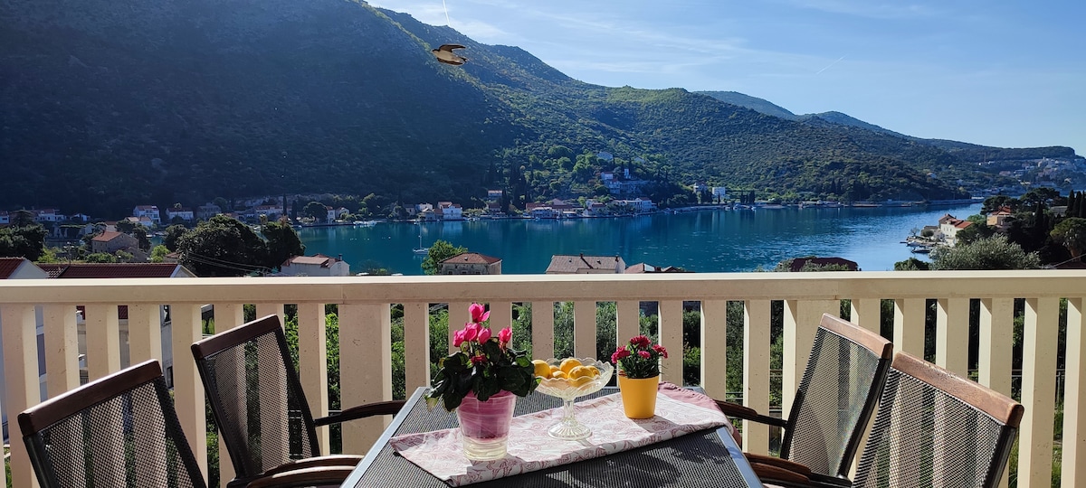 A balcony table is set with potted flowers and a bowl of fruit, overlooking a serene sea view framed by rolling hills. Four chairs surround the table, and the bright morning light enhances the natural colors of the scene.
