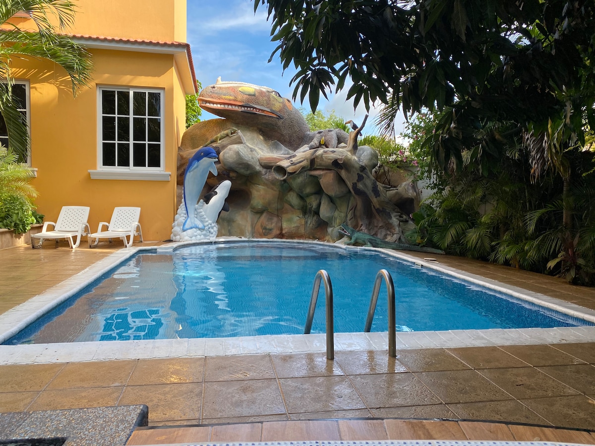 A sparkling pool is framed by tropical plants and a decorative rock feature. Two lounge chairs are positioned nearby, inviting relaxation. Sunlight reflects off the water's surface, creating a serene setting for leisure or family time.
