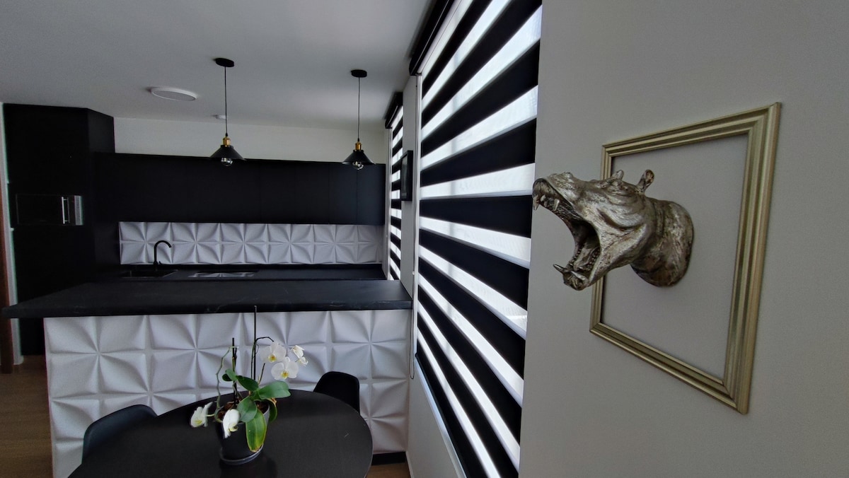 An integrated kitchen area showcases a sleek, black countertop and modern cabinetry, with unique paneling. Black and white striped window treatments allow natural light to filter in. A decorative wall mount in the shape of a hippo adds an artistic touch, while a small table features a floral arrangement.