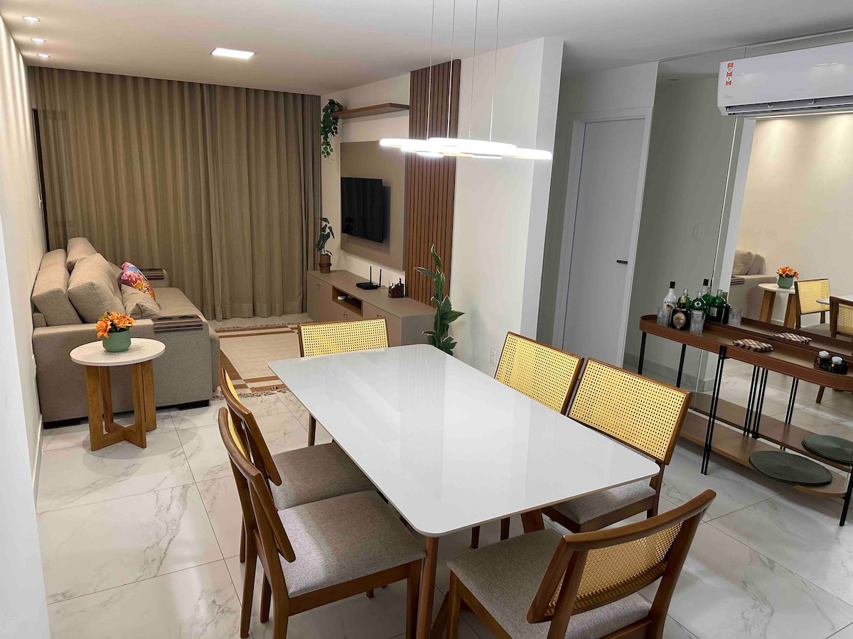 A spacious living and dining area is visible, featuring a modern dining table with eight chairs and a cozy sofa across the room. Soft, neutral tones are complemented by warm lighting, and a television is mounted on the wall. Decorative plants add a touch of greenery.