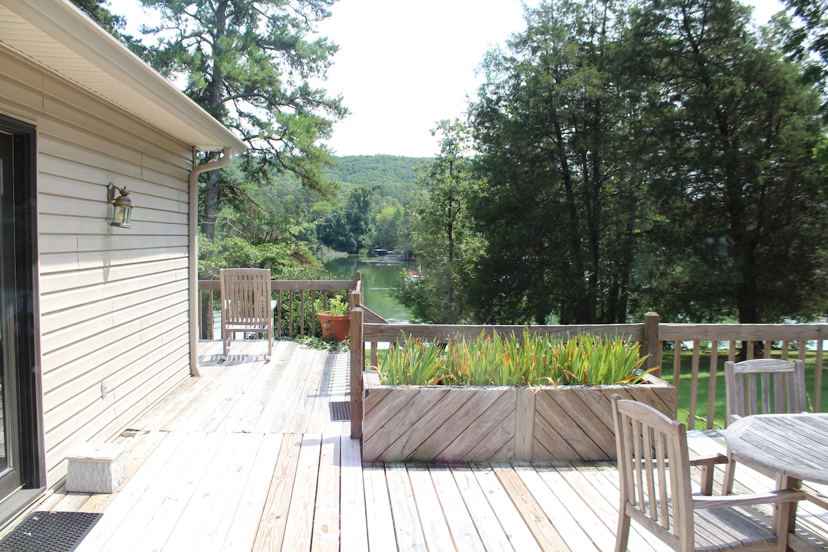 A wooden deck features a spacious seating area with two chairs positioned for lake views. A raised planter filled with greenery adds a natural touch. Surrounding trees provide shade and privacy, while the serene lake can be glimpsed in the background.