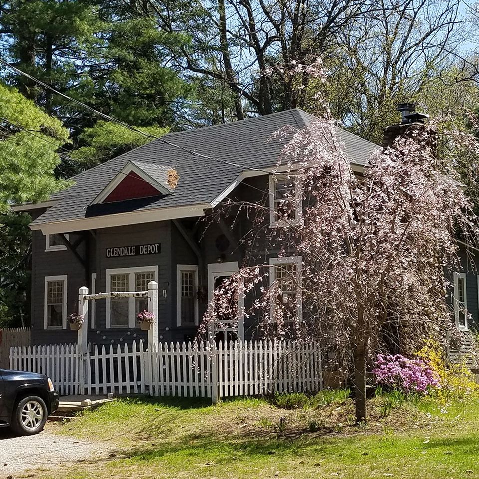 The charming exterior of the Train Station House is showcased, featuring a grey facade and white picket fence. Delicate pink blossoms are visible on a nearby tree, with vibrant flowers blooming in the yard. A driveway leads towards the house, enveloped by lush greenery.