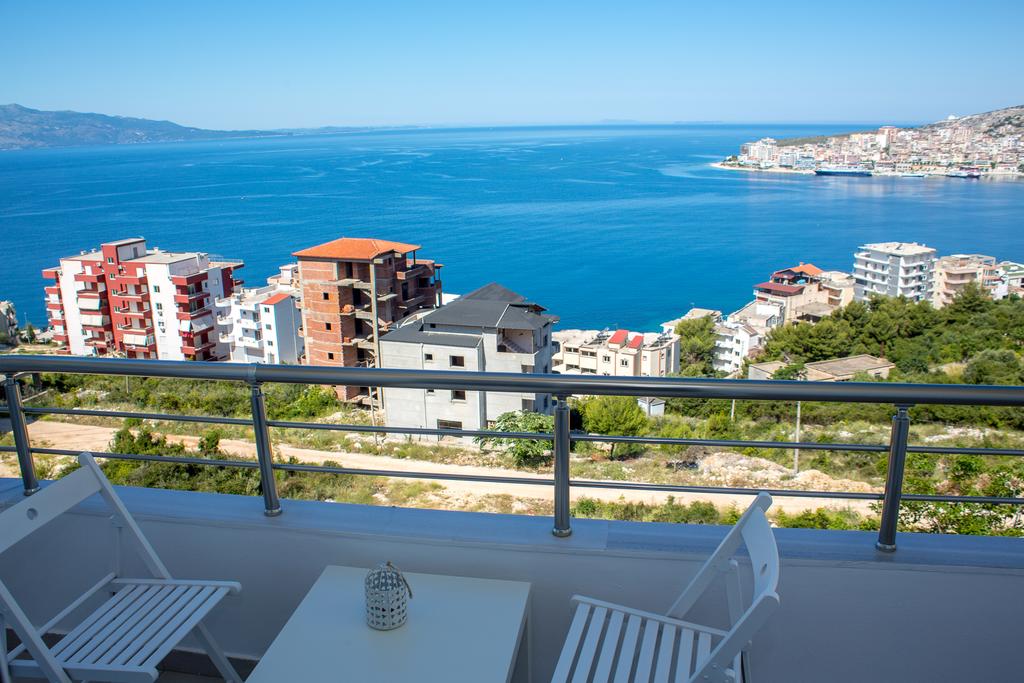 A balcony offers stunning views of the coastline and the sea, accompanied by a small white table and two white chairs. The scenery includes buildings arranged along the waterfront, with mountains visible in the distance under a clear blue sky.